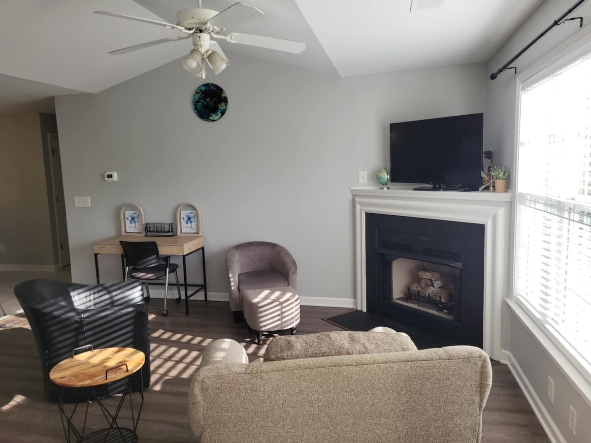 A comfortable living area features a gray sofa, a small wooden table, and two armchairs. A ceiling fan provides airflow above, while natural light streams through large windows. A modern fireplace and a television are positioned on the wall, enhancing the relaxed atmosphere.