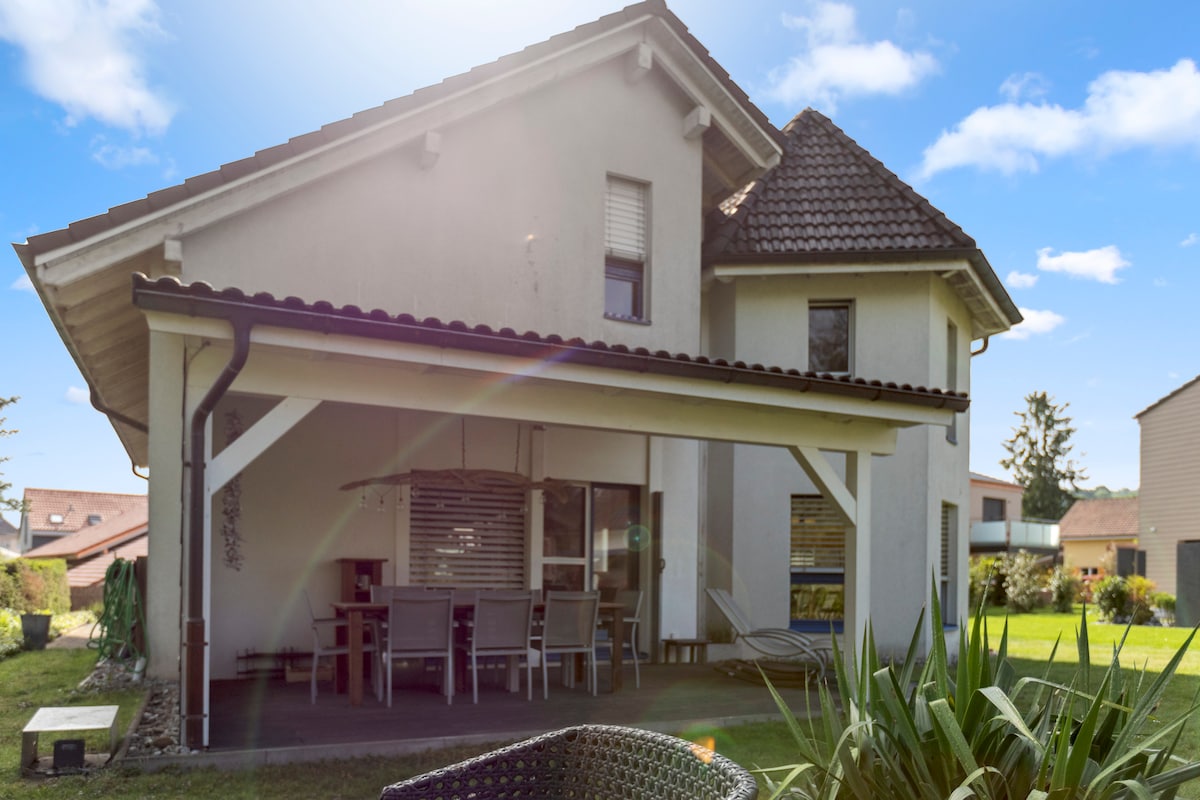 The exterior of the house features a covered terrace with a dining area, surrounded by a lush garden. The structure highlights a sloped roof and large windows, allowing natural light to fill the space. A patio is visible at the base of the garden.