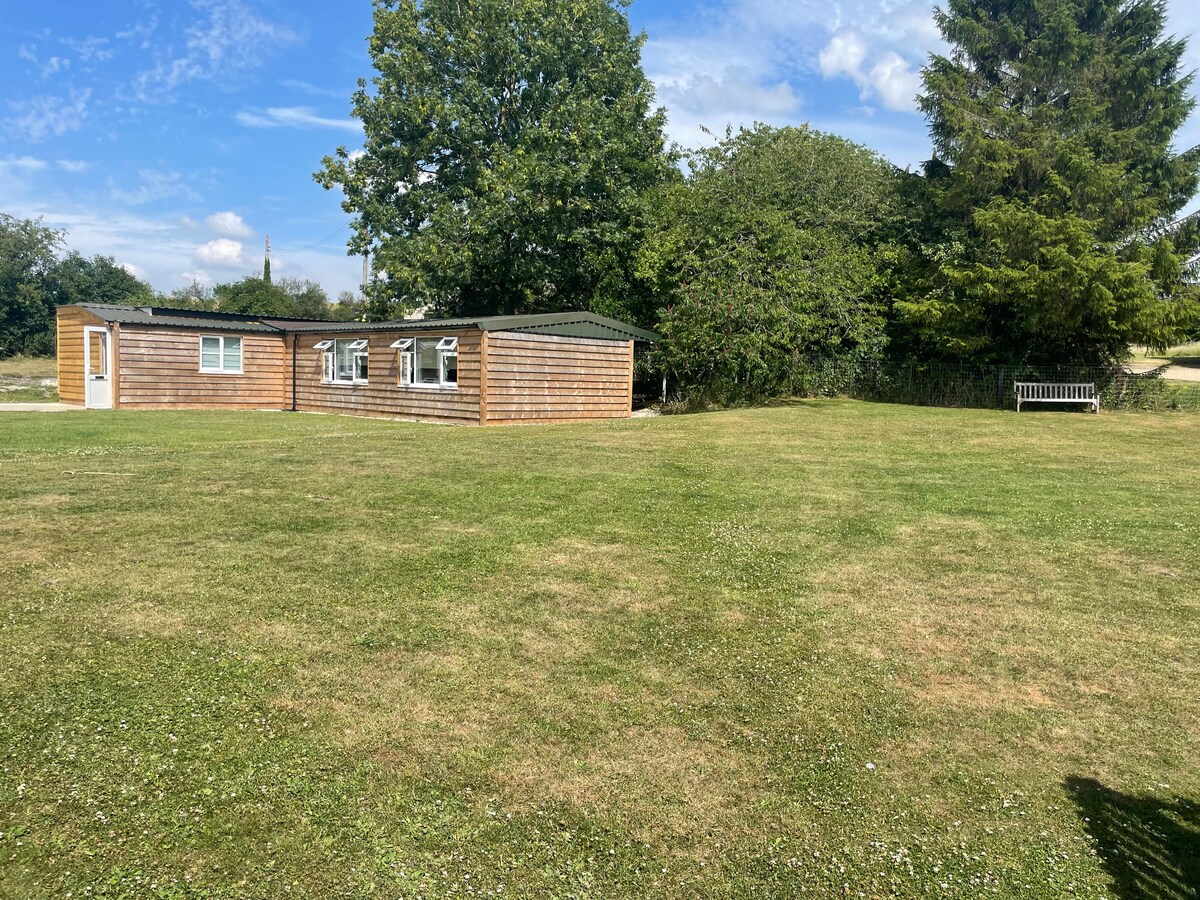 The timber cabin is positioned on a spacious green lawn, surrounded by trees and shrubs. A bench sits to the right, offering a place to relax. The sky is clear with scattered clouds, enhancing the sense of openness in the natural setting.