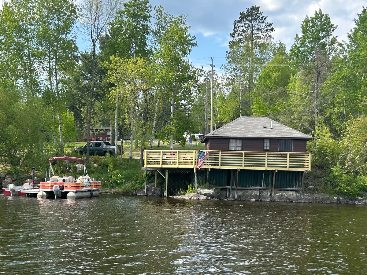 A cabin is situated by the water's edge, surrounded by lush greenery. A wooden deck extends along the exterior, and a flag is displayed prominently. A boat is docked nearby, showcasing the peaceful lakeside setting.