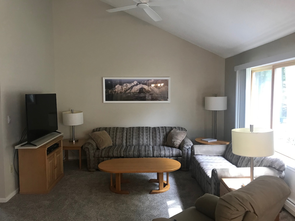 A comfortable living area features a striped sofa and a matching loveseat, complemented by light wood tables. A flat-screen television is positioned on a wooden console. Natural light filters through large windows, adding brightness to the space.