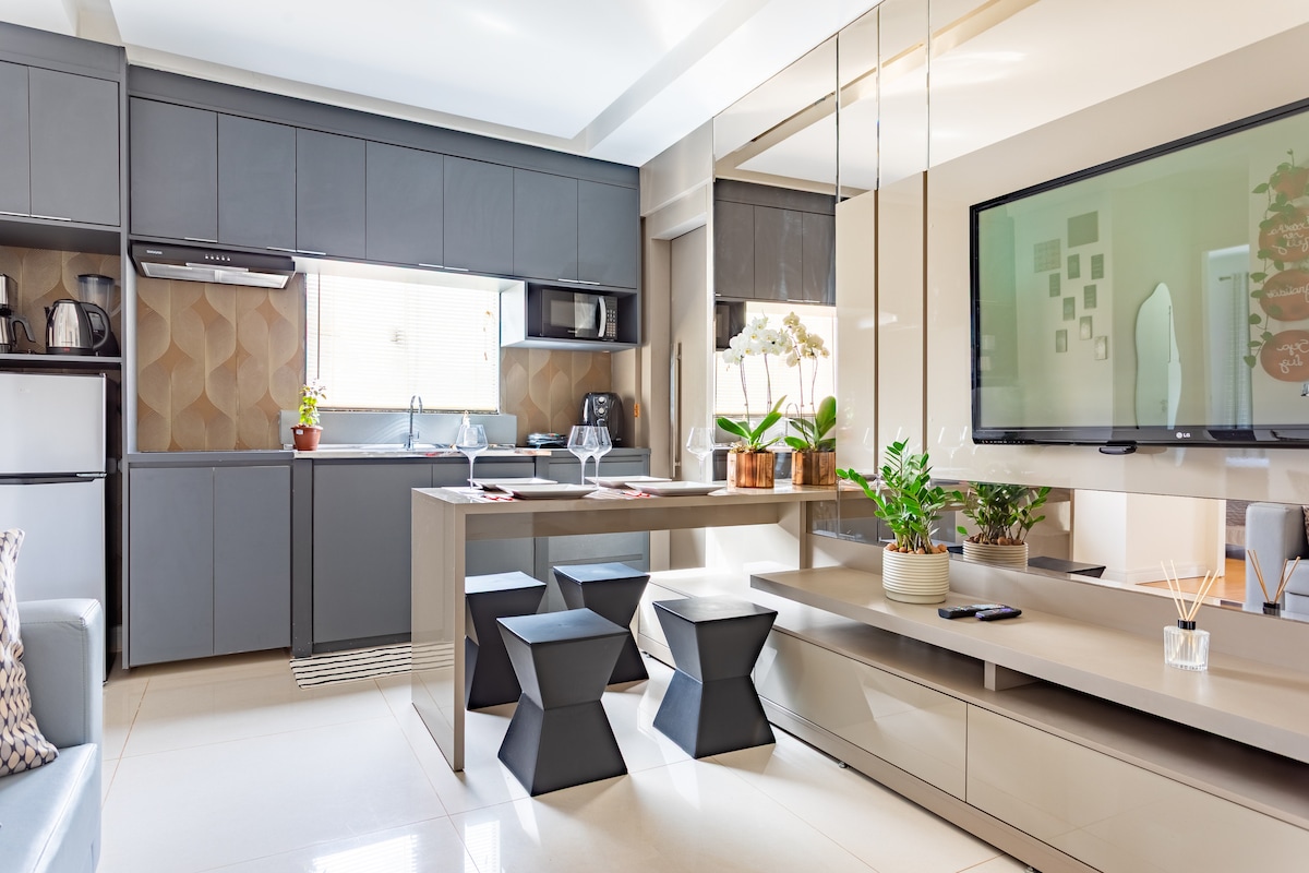 A modern kitchen and dining area features a functional layout with sleek gray cabinetry and a mirrored wall. A dining table is positioned at the center, surrounded by black geometric stools. A large TV is mounted on the wall, enhancing the spacious feel of the room.