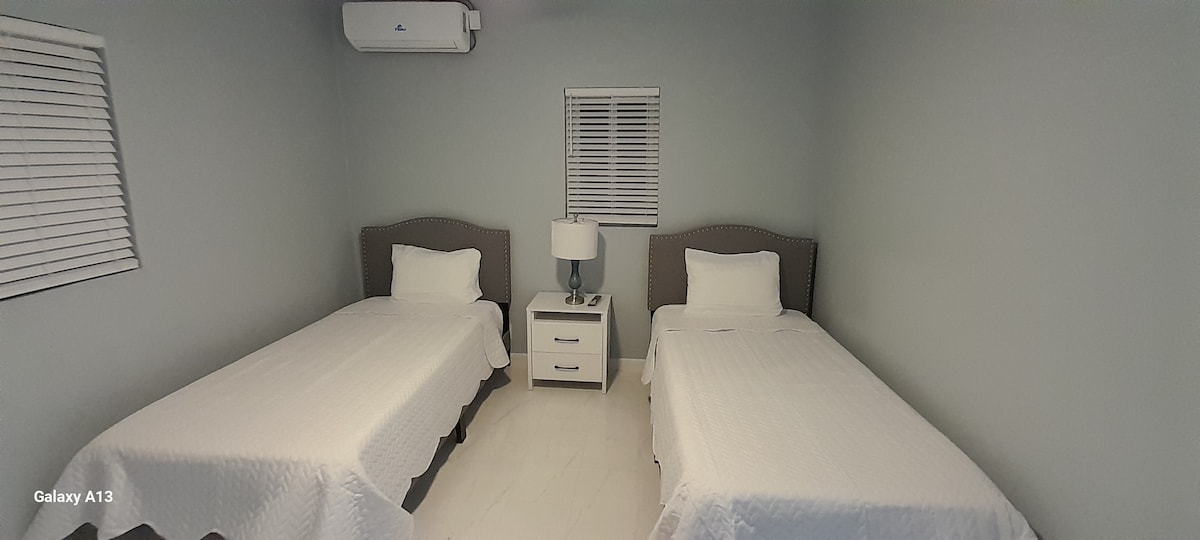 The sleeping area contains two twin-sized beds with decorative headboards, neatly covered with white linens. A nightstand with a lamp is positioned between the beds, and a small window is dressed with white blinds, allowing soft light to filter into the room.