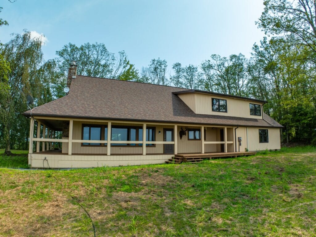 The exterior of the spacious retreat features a two-story structure with a sloped roof and multiple windows. A wrap-around porch provides access to the outdoors, surrounded by greenery. The setting is peaceful, with trees lining the background and a natural grass area in the foreground.