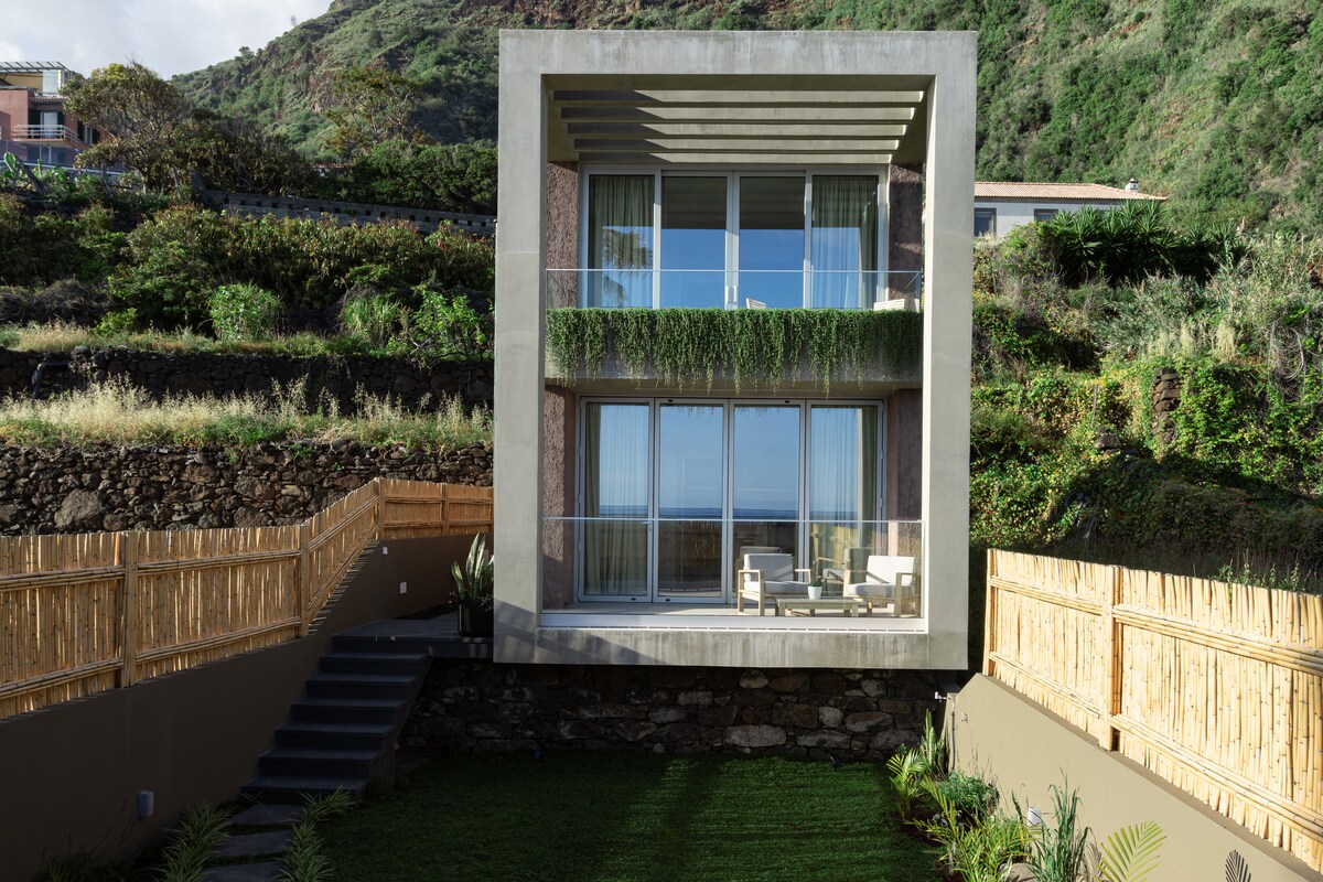 A modern building is set against a lush green hillside, featuring large windows that provide natural light and views. A small garden area with grass is visible, alongside a wooden fence that enhances the property’s spacious feel.