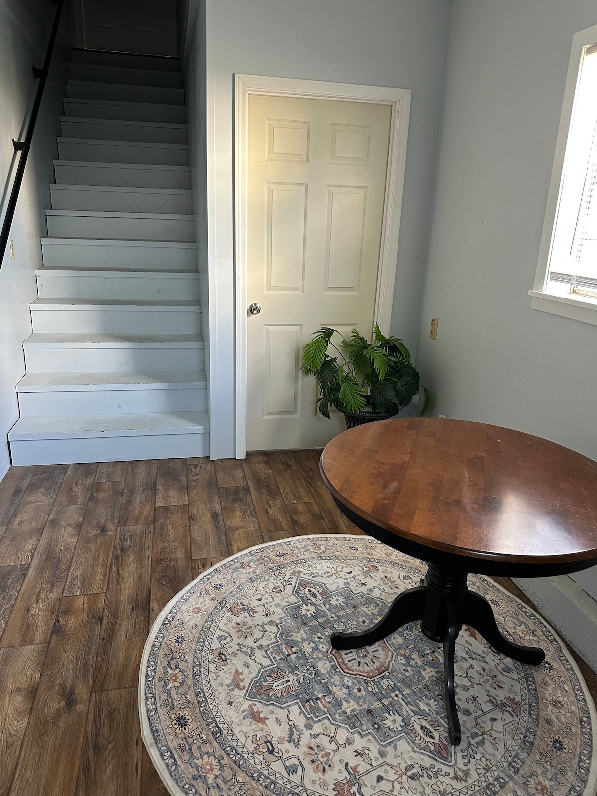 A welcoming entryway features a round wooden table situated on a decorative circular rug. Light streams in through a nearby window, illuminating the space. A set of stairs leads to an upper level, while a green plant adds a touch of nature to the corner.