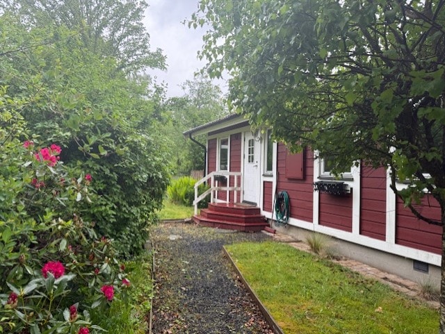A charming exterior is framed by lush greenery and blooming flowers. A stone pathway leads to a welcoming entrance with a red and white facade. The surrounding landscape features various bushes and trees, creating a serene environment.