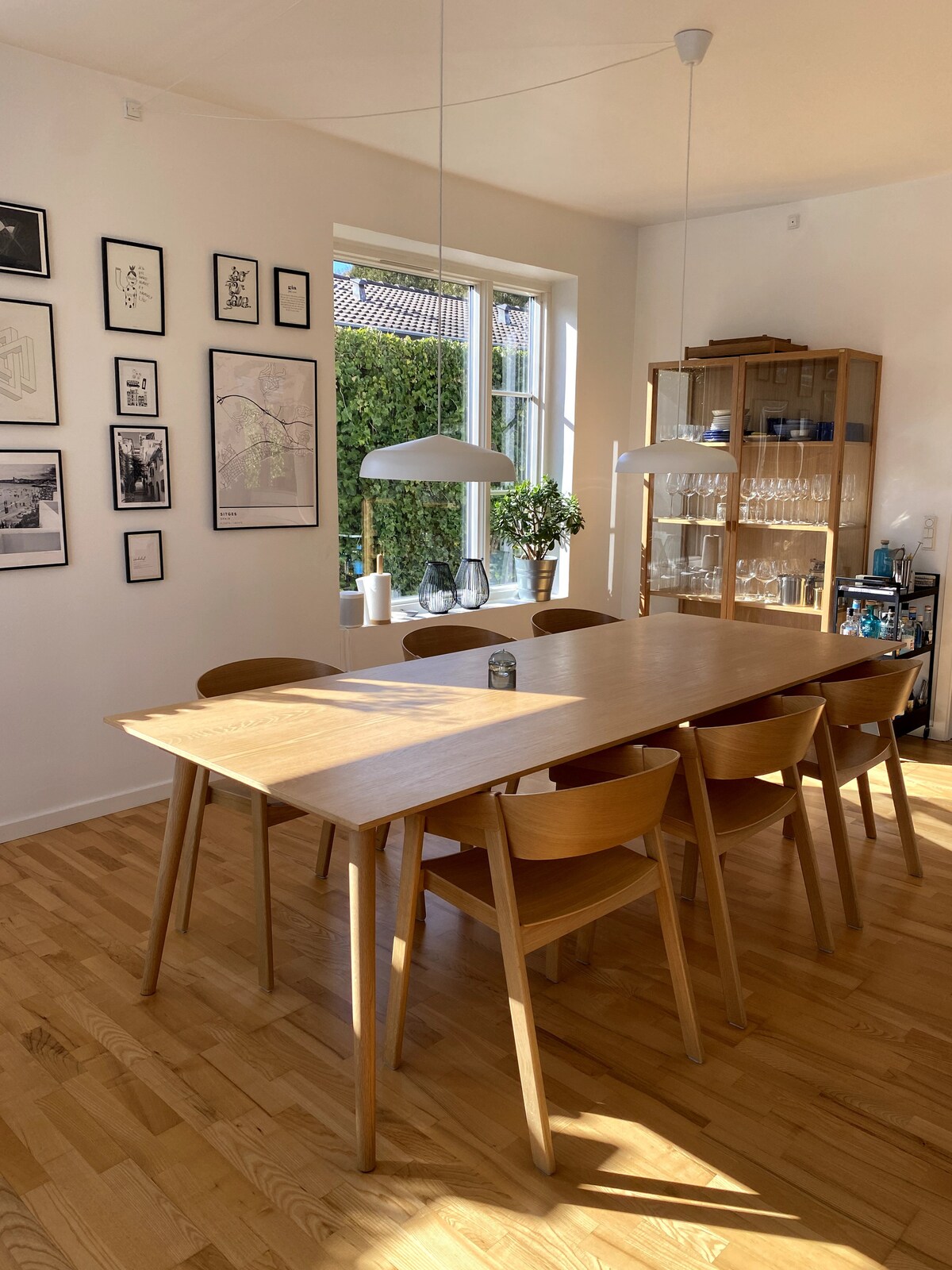 A dining area features a wooden table surrounded by six chairs, illuminated by soft natural light from a large window. The walls are adorned with various framed artworks. A glass cabinet holds neatly arranged glassware, contributing to the clean and modern aesthetic.