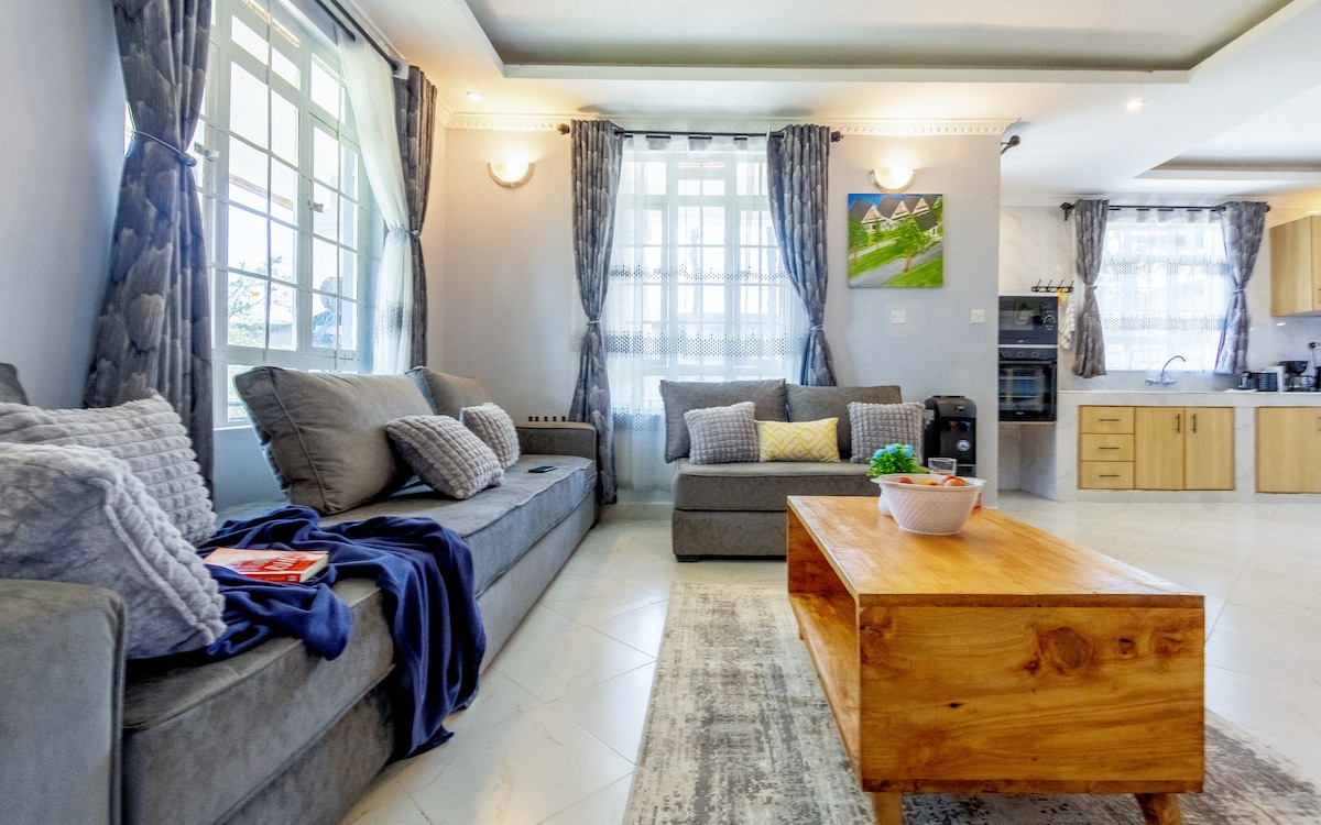 A spacious living area features plush grey sofas accented with a variety of cushions and a blue throw. A wooden coffee table sits at the center, with natural light filtering through large windows adorned with elegant curtains.