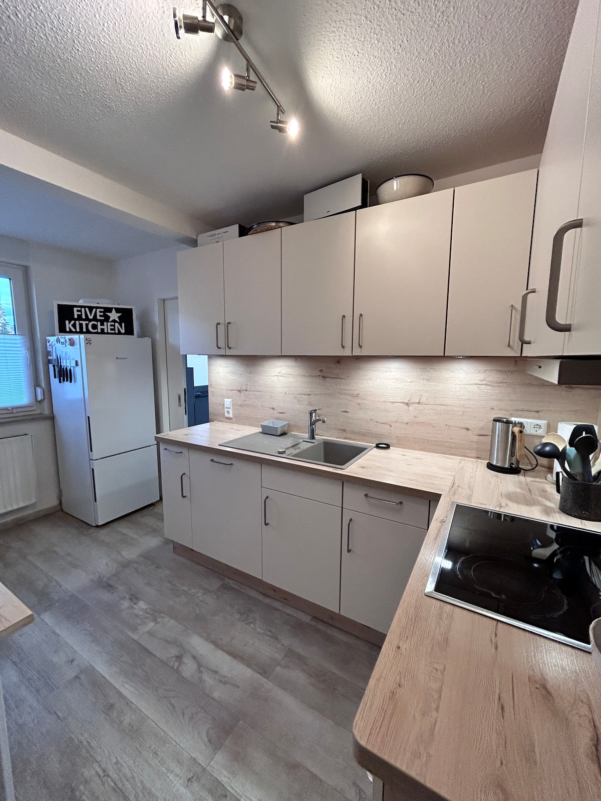 The kitchen features modern cabinetry and a light wood backsplash. Stainless steel appliances, including a fridge and kettle, are visible. Under-cabinet lighting highlights the countertops, while a sink and cooktop are adequately placed for functionality.