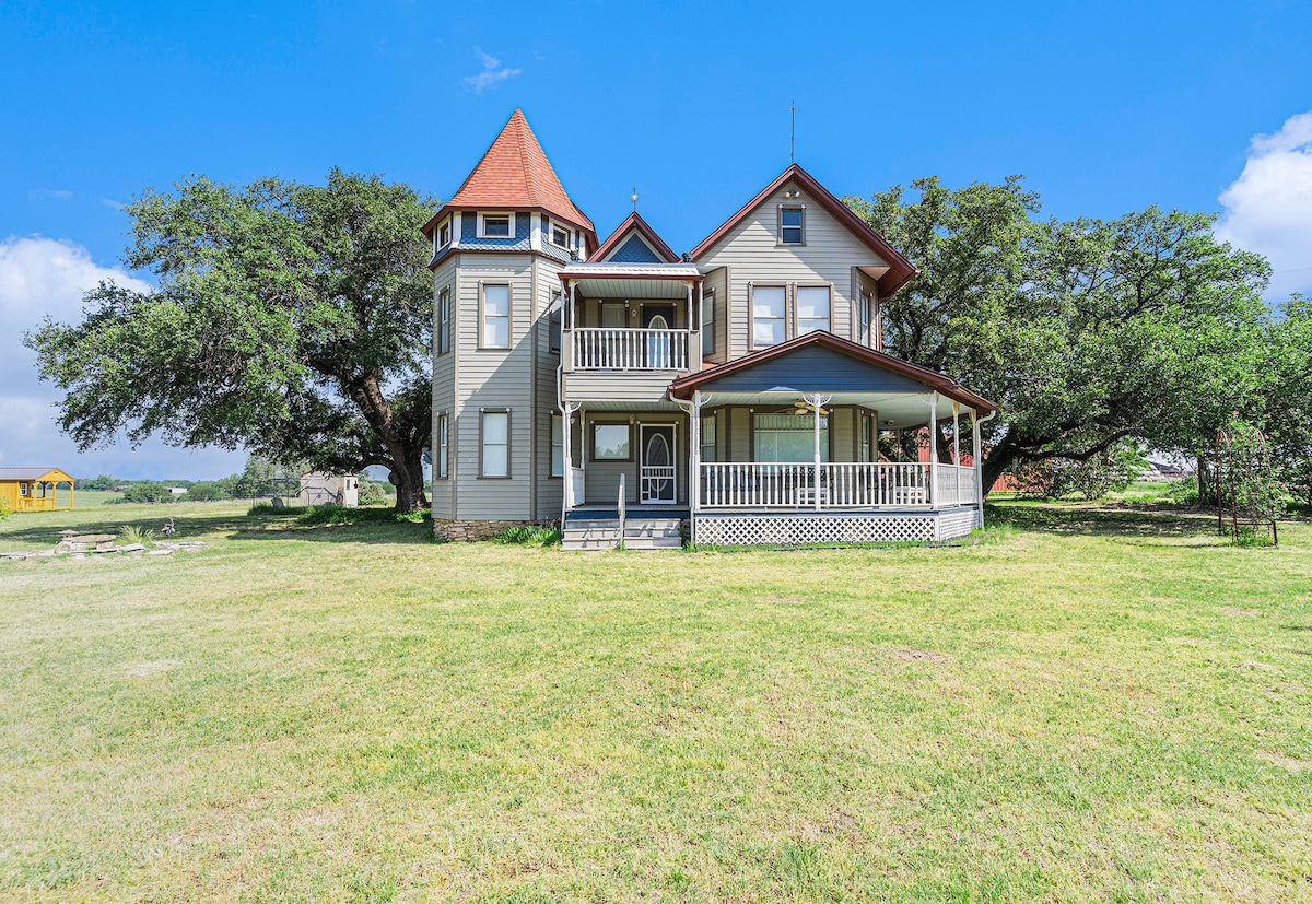 A three-story Victorian country home is set against a backdrop of rolling pastures. The structure features a turret with a pointed roof, a welcoming porch with a railing, and expansive green lawn that invites outdoor activities.