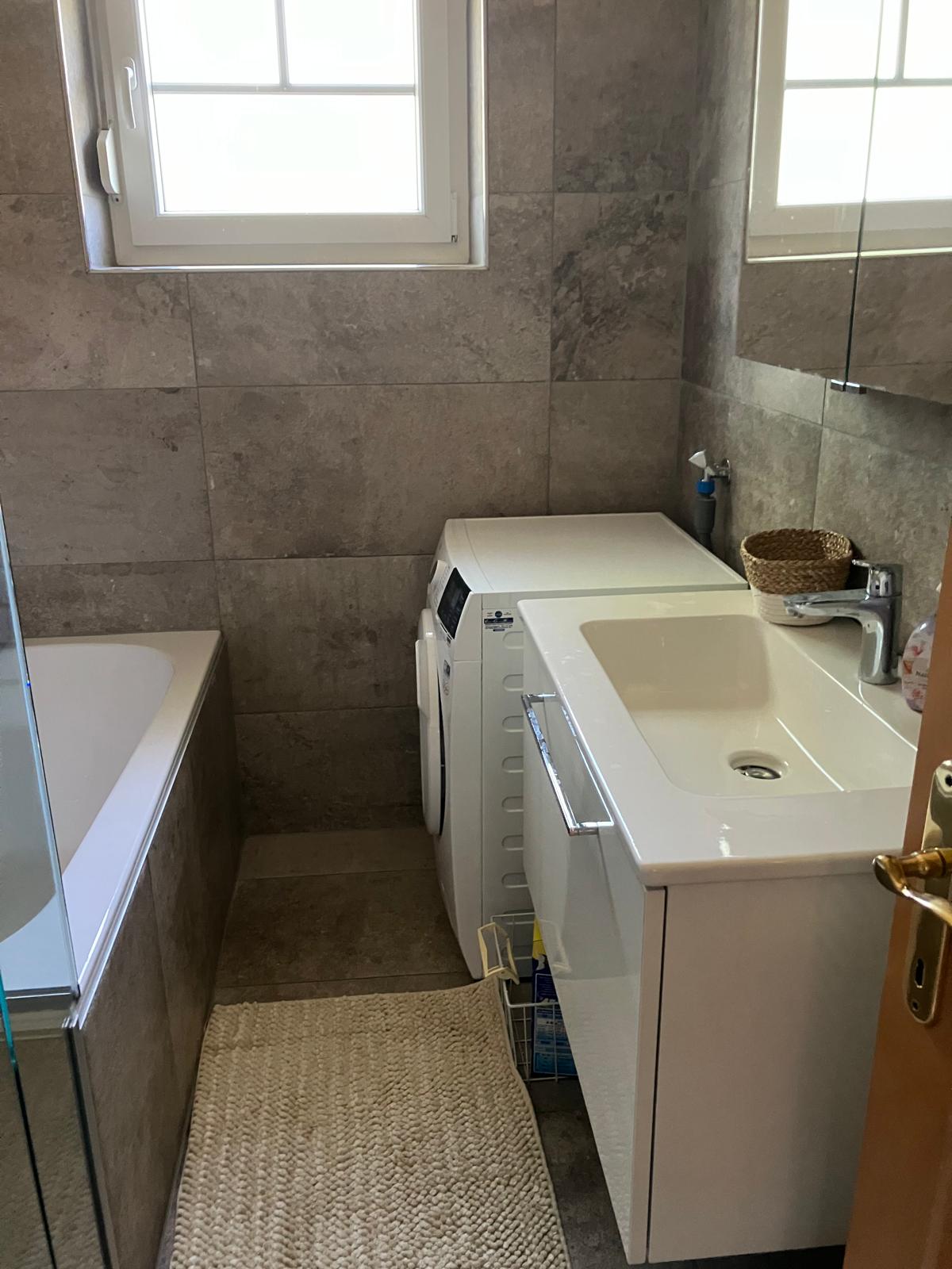 A modern bathroom is presented, featuring a sleek white sink and a washer integrated into cabinetry. A glass shower enclosure and a bathtub are visible, complemented by neutral-toned tile walls. Natural light enters through a nearby window, enhancing the clean and functional design.