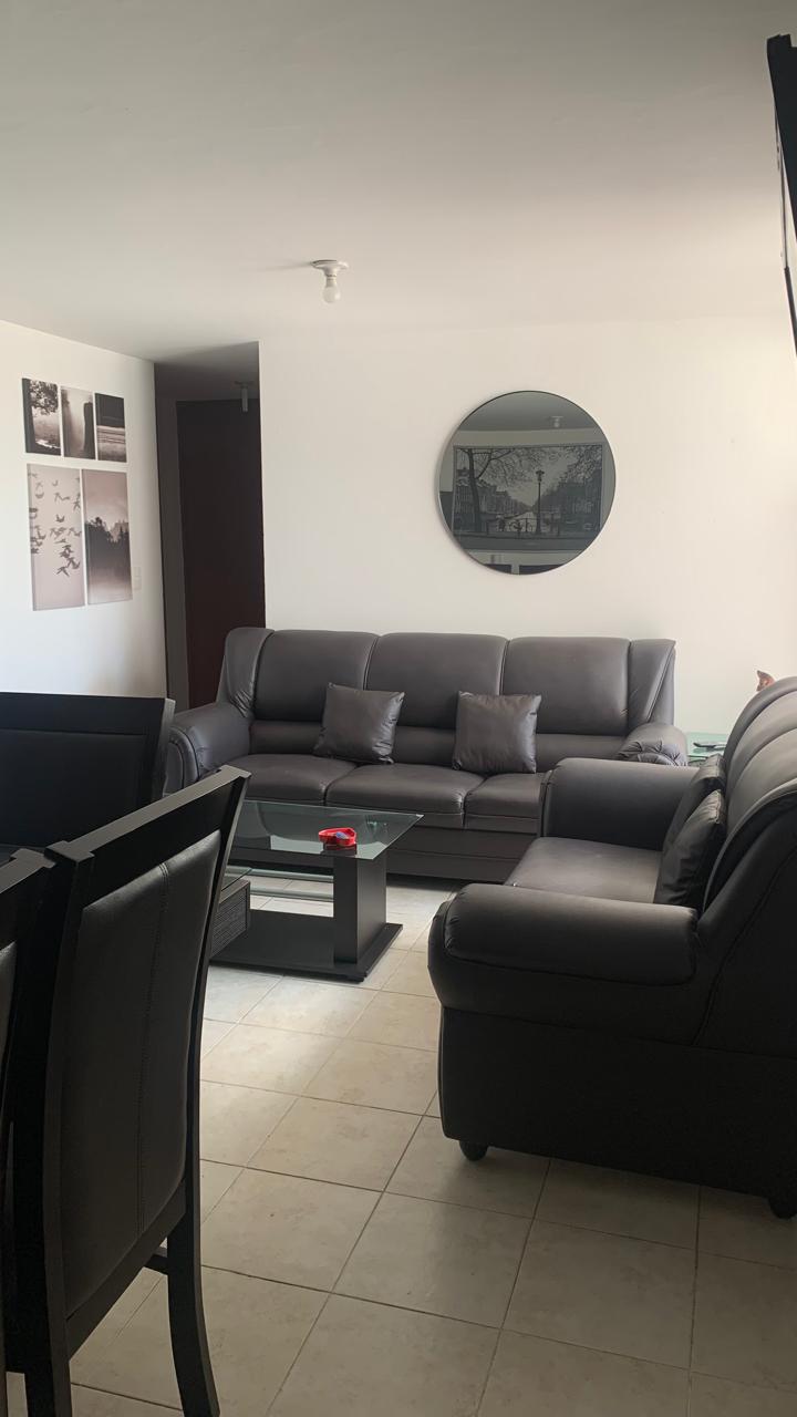 A cozy living area features a dark leather sectional sofa with several cushions. A small glass coffee table sits in front, complementing the low dining table seen in the background. Monochrome wall art adds interest to the space, highlighted by natural light from a window.
