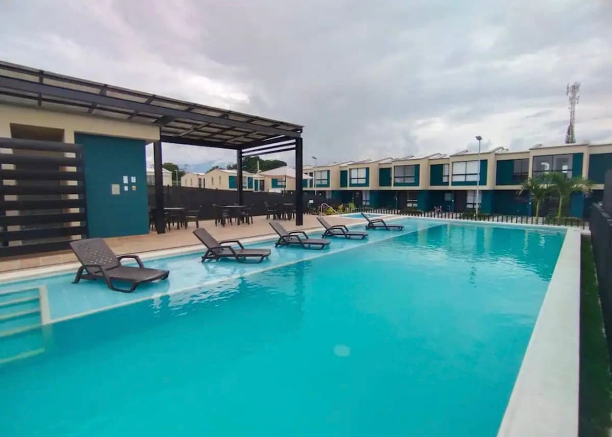 A serene swimming pool is framed by a sun deck featuring several lounge chairs. The pool reflects a calm blue, with a covered seating area nearby. Two-story buildings in the background create a balanced view, complemented by a lush green landscape.