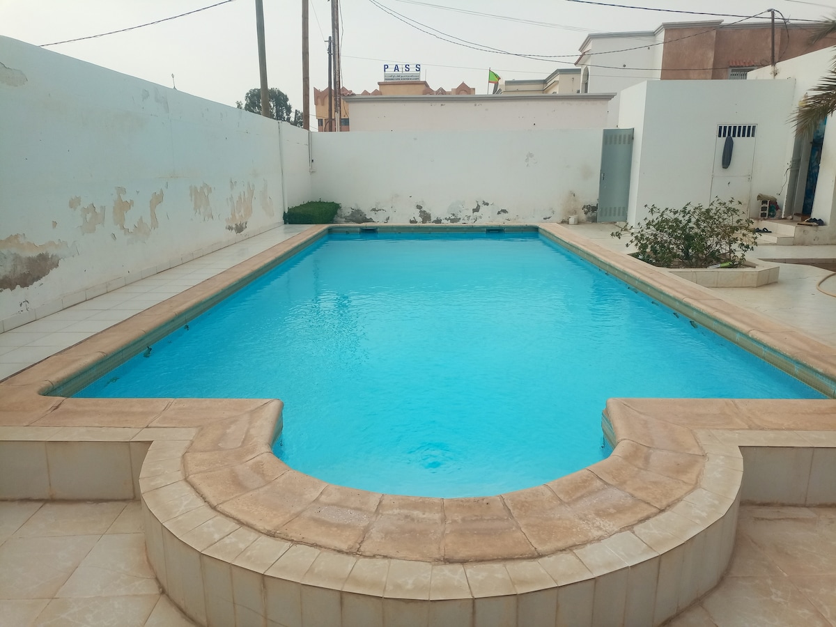 A clear swimming pool is featured in a private courtyard, surrounded by tiled flooring and a simple white wall. Lush green plants are positioned at one end of the pool, while the sky is visible above, showcasing a hint of urban surroundings.