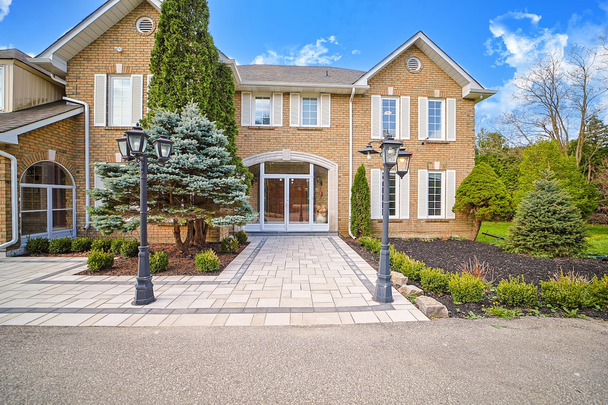 An elegant brick façade is presented, framed by well-manicured shrubs and trees. A spacious walkway made of light-colored pavers leads to a central entrance, flanked by decorative lanterns. Clear blue skies and gentle greenery enhance the outdoor area's inviting appeal.
