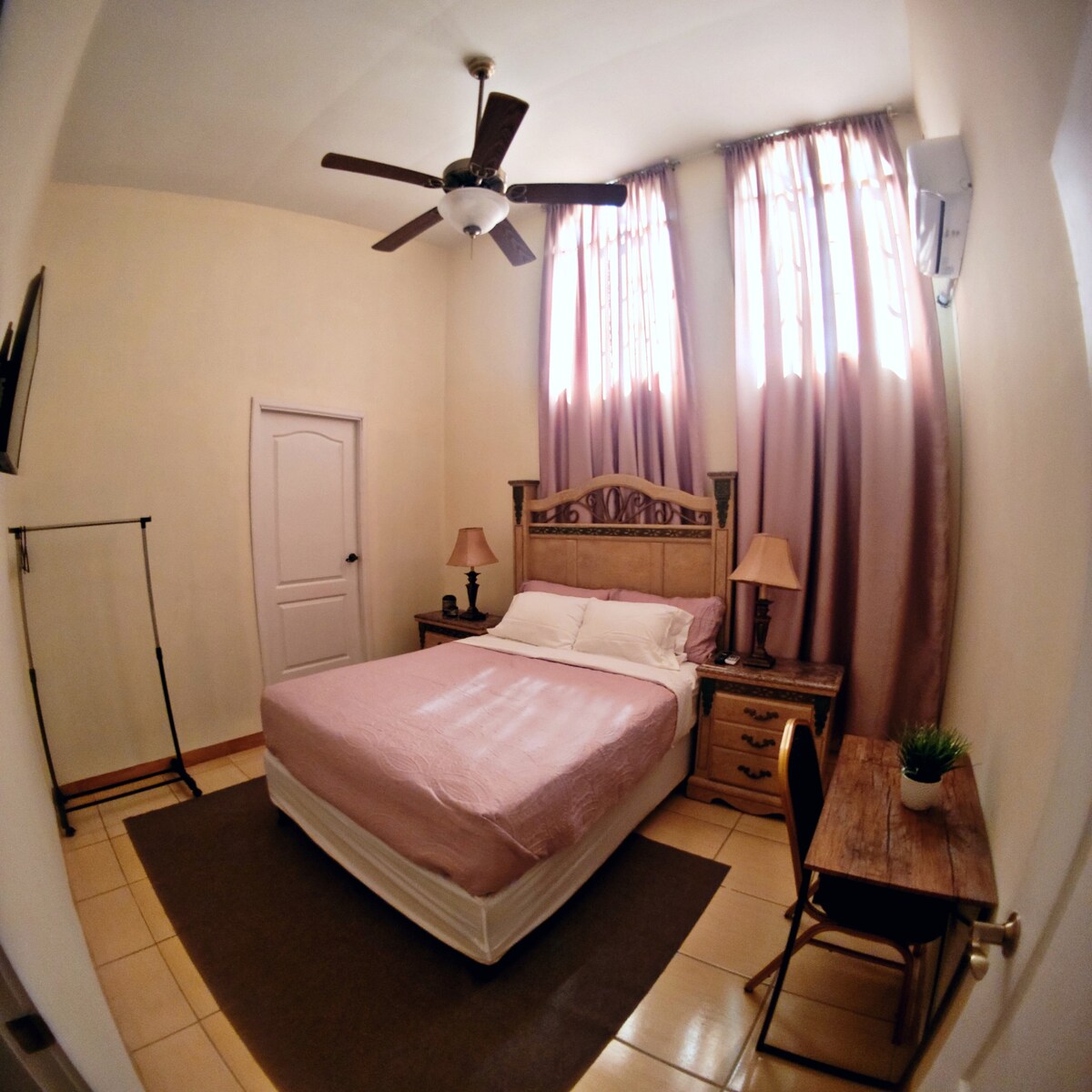 A comfortable bedroom features a spacious bed with light bedding. Two bedside lamps are placed beside the bed, and natural light filters through tall windows dressed with soft curtains. A small desk and chair sit against one wall, providing a cozy workspace.