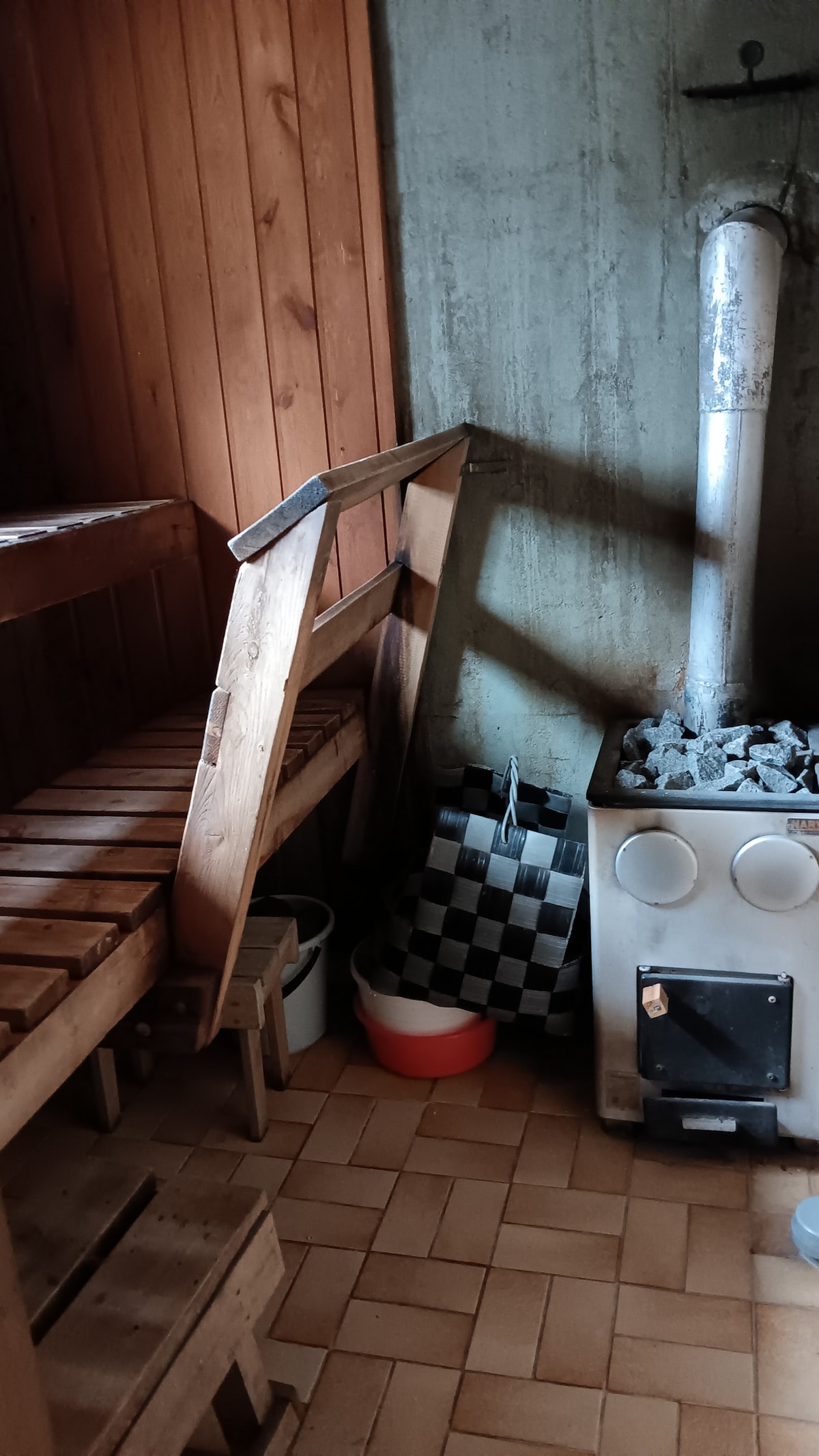 A sauna area is shown with wooden benches against a smooth, stone wall. A metal stove with visible stones sits in the corner, providing warmth. Additional items, including buckets and checkered towels, are placed nearby on the tiled floor.