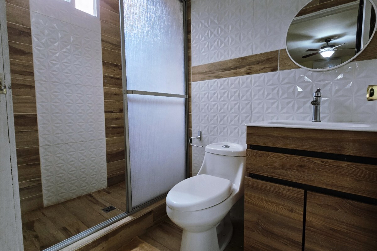 A modern bathroom features a walk-in shower enclosed by a glass partition. A circular mirror reflects soft lighting above a wooden vanity with a sink. A toilet is positioned conveniently nearby, complemented by textured wall tiles and a warm wooden floor.