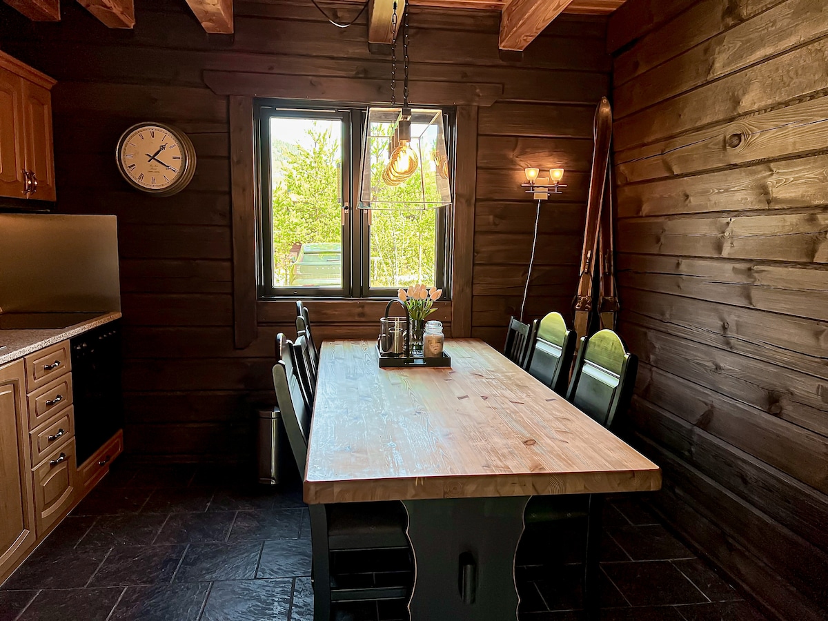 A rustic dining area features a wooden table surrounded by black chairs, positioned beneath warm lighting. A large window provides a view of the outdoors, while wooden walls enhance the cozy atmosphere. An elegant clock and vintage ski equipment are displayed for added charm.