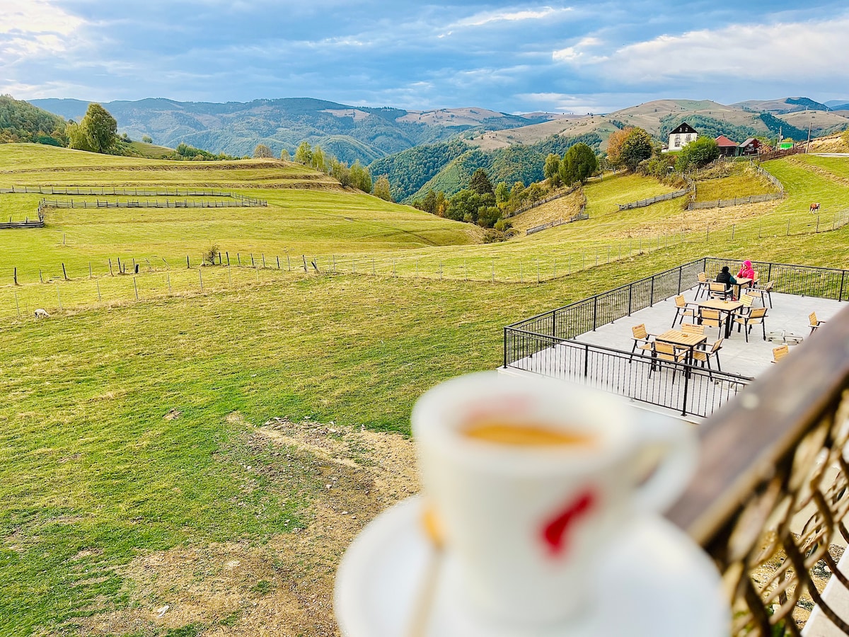 A view of rolling green hills stretches across the landscape, creating a serene backdrop. In the foreground, a cup of coffee is held at the edge of a balcony, while a patio area with tables and chairs is visible, suggesting a peaceful outdoor space for relaxation.