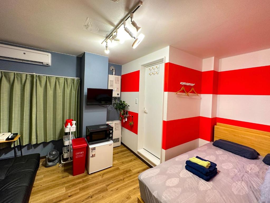 A studio space is divided by red-and-white striped walls, offering a cozy sleeping area with a double bed covered in linens. A compact kitchenette is visible, featuring a microwave and refrigerator. Natural light filters through green curtains, adding warmth to the environment.