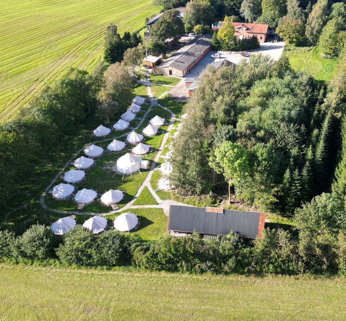 An aerial view captures a serene landscape featuring multiple white tents arranged in a circular pattern. Surrounding greenery and trees provide a natural setting, while a cluster of buildings is visible in the background, offering additional facilities and accommodations.