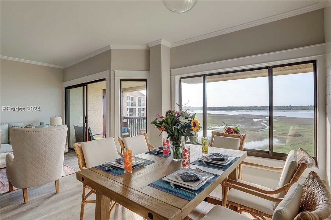 A spacious dining area is designed with a wooden table set for six, featuring colorful table decor and flowers. Large windows frame views of the marsh, inviting natural light into the space, while a cozy seating area can be glimpsed nearby.