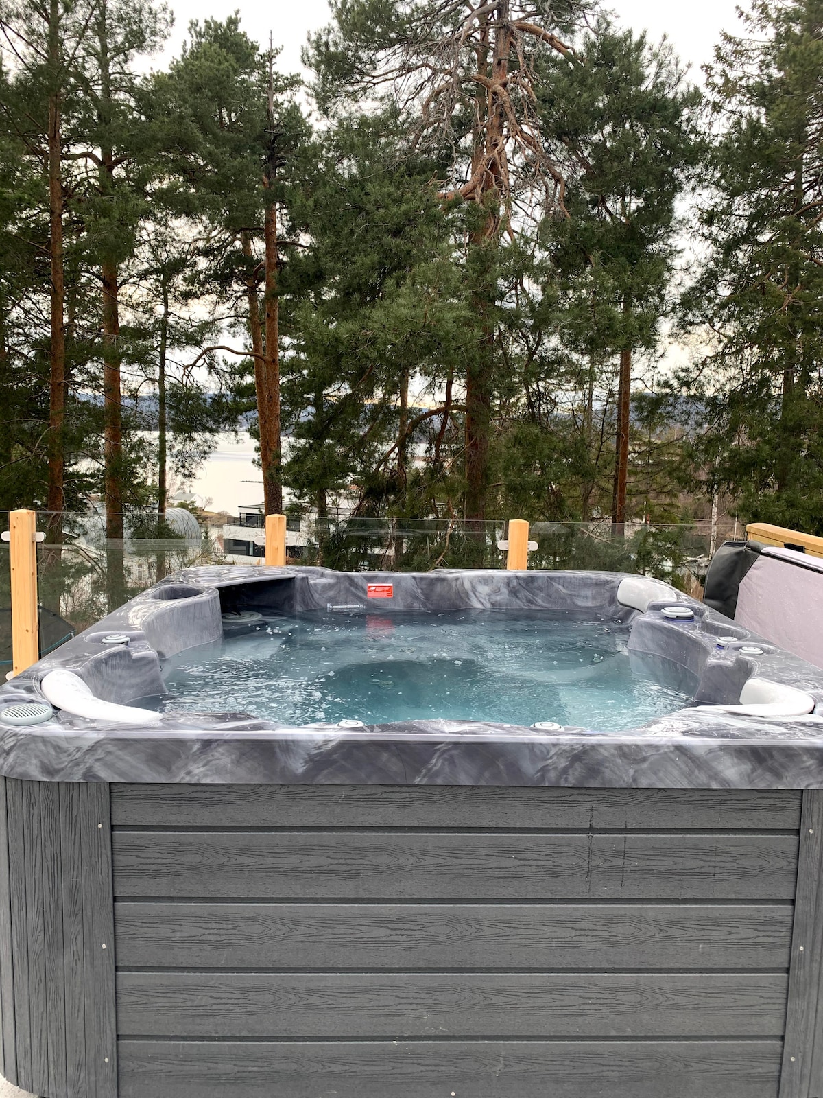 A private jacuzzi is situated on a terrace, surrounded by tall trees. Bubbles rise from the water, creating a relaxing atmosphere. Clear glass panels offer an unobstructed view of the surroundings, enhancing the tranquil environment.