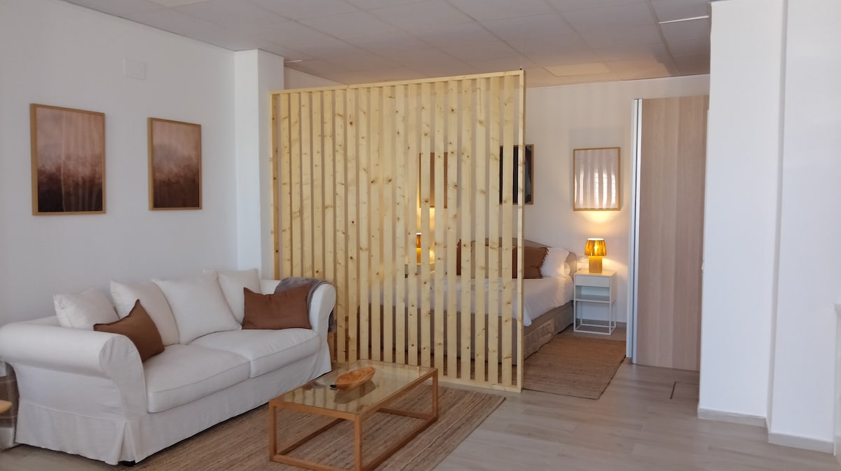 An open living area features a light-colored sofa with decorative pillows, complemented by a wooden coffee table. Behind a wooden partition, a cozy sleeping area is visible, furnished with a spacious bed and warm lighting. Neutral tones and wall art enhance the inviting ambiance.