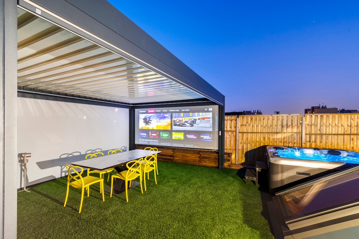 An outdoor dining area features a long table surrounded by six bright chairs, set within a covered pergola. A large projection screen displays various options for entertainment, while a hot tub is located nearby, providing a cozy atmosphere for relaxation under the evening sky.