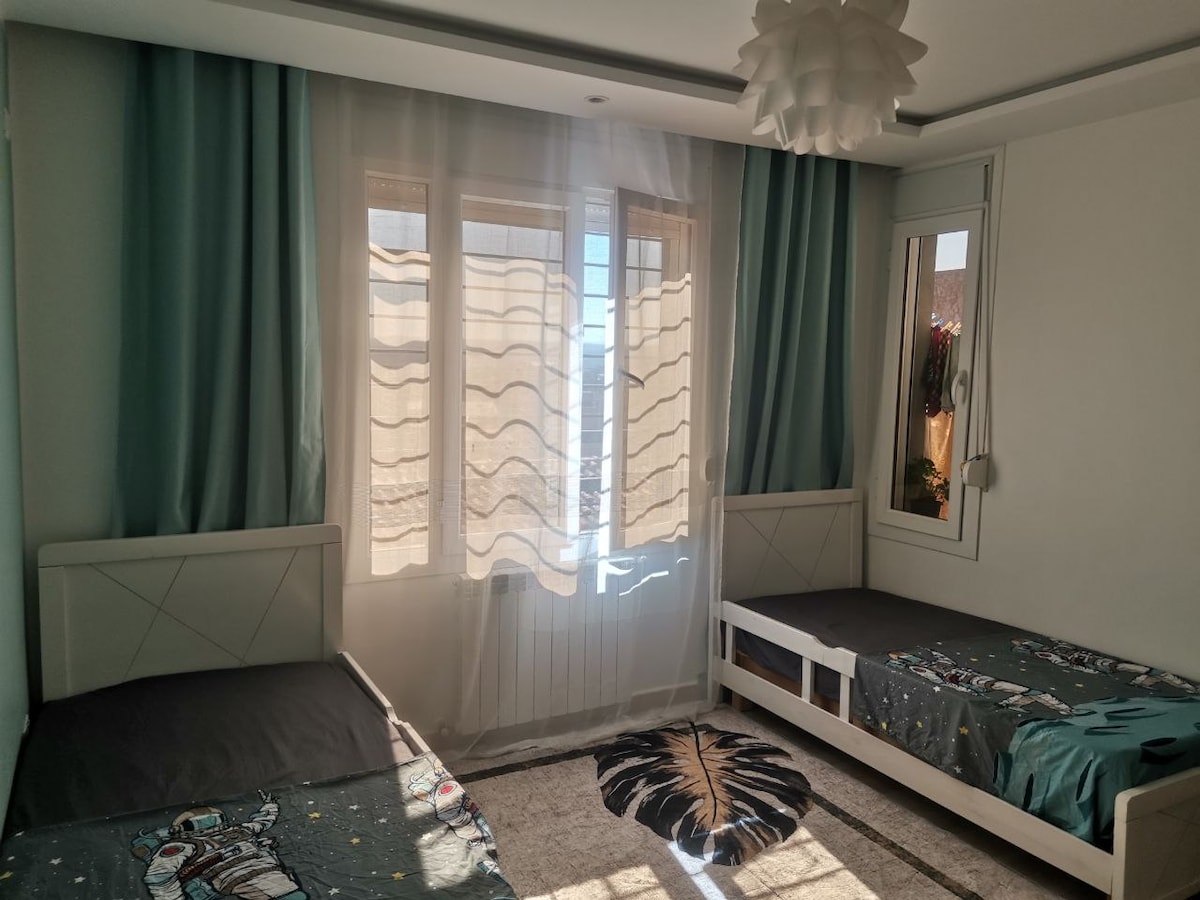 A well-lit sleeping area features two separate beds with dark bedding. Sheer curtains gently filter light, casting soft shadows across the room. A decorative rug with a leaf pattern lies on the floor, complementing the light green walls and providing a cozy touch.