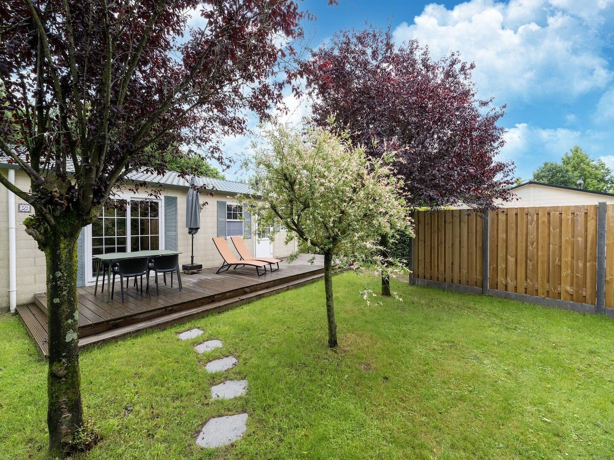 A serene outdoor space is shown, featuring a wooden deck with two lounge chairs. Lush greenery surrounds the area, including a flowering tree and vibrant foliage. A fenced boundary provides privacy, while a stone path leads through the well-kept lawn.
