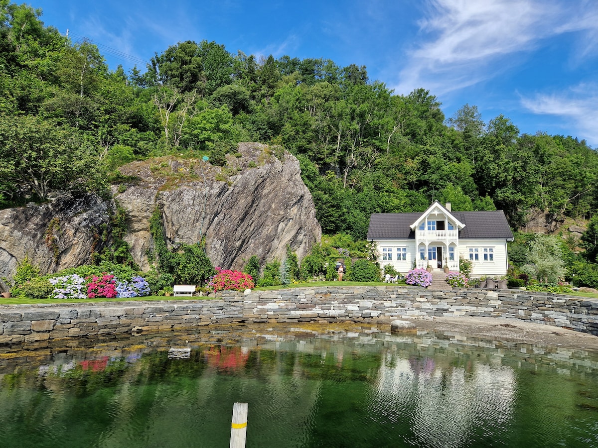A modern villa with large windows and a welcoming entrance is set against a backdrop of lush greenery and rocky hills. Colorful flowerbeds in bloom line the shore, reflecting beautifully in the calm water, creating a serene and inviting outdoor space.