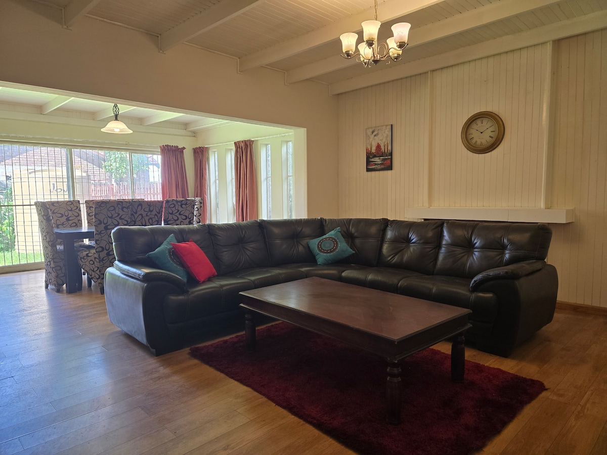 A spacious living area features a dark leather sectional sofa adorned with colorful cushions. A wooden coffee table is positioned in front of the sofa. Large windows with sheer curtains allow natural light to fill the room, highlighting the wooden floor and decorative wall clock.