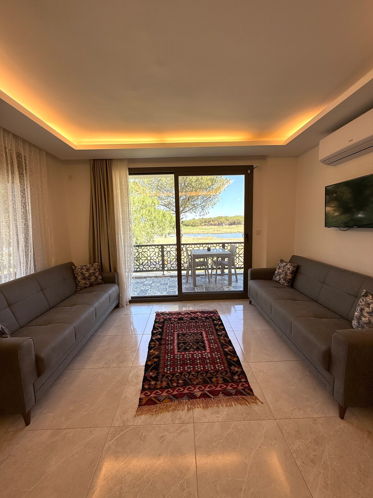 A bright living area is depicted, featuring two gray sofas positioned opposite each other. A decorative rug lies centered on the glossy tile floor. Large glass doors offer views of the outdoors and lead to the balcony, while light-colored curtains frame the windows.