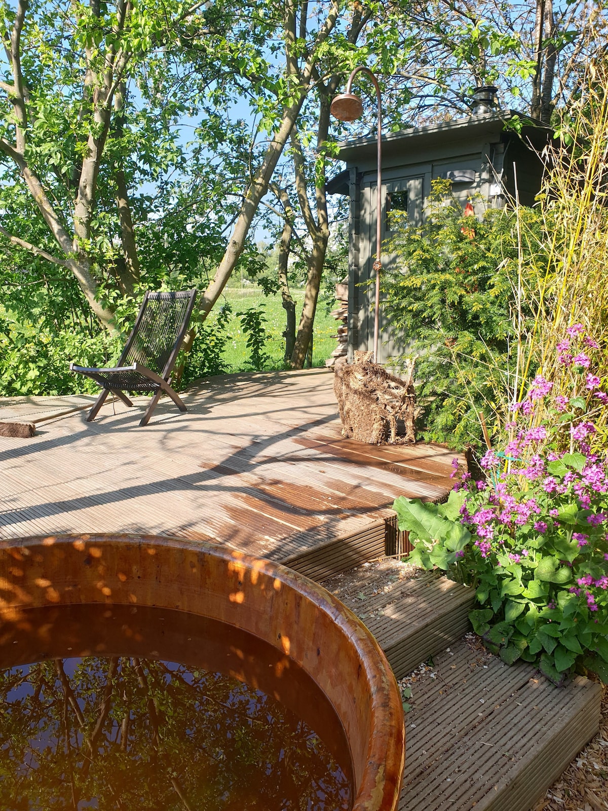 A wooden deck area features a rustic hot tub surrounded by lush greenery and vibrant flowers. A chair is positioned nearby, providing a place to relax in the serene outdoor setting, with a glimpse of the garden and a small structure in the background.