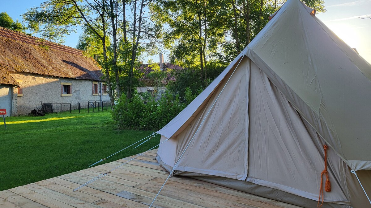 A spacious tent is set on a wooden deck, surrounded by lush green grass and trees. In the background, a rustic building with a thatched roof is visible, offering a serene outdoor environment. Soft natural light enhances the tranquil atmosphere.
