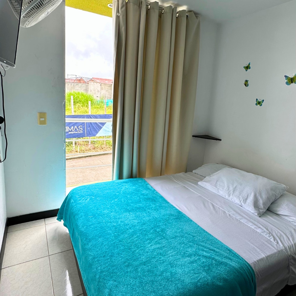A cozy bedroom features a double bed covered with a white sheet and a turquoise throw, complemented by soft natural light filtering through the curtains. A small shelf beside the bed holds minimal decor, and butterfly wall art adds a subtle touch of color.