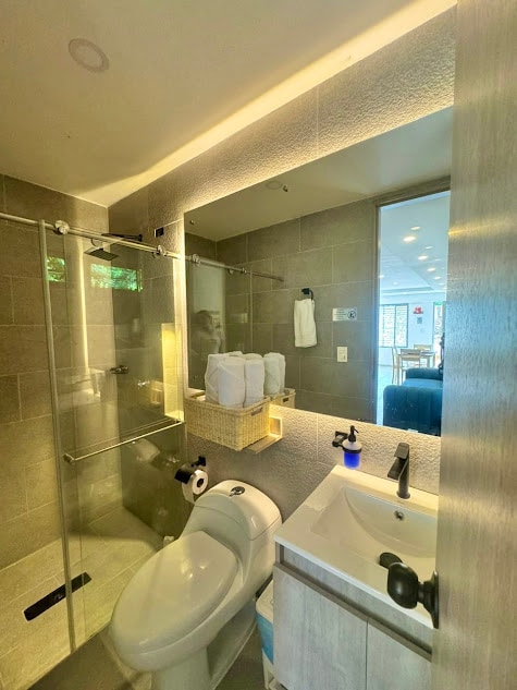 A modern bathroom is depicted, featuring a walk-in glass shower with a rainfall showerhead. A sleek vanity with a small basin is complemented by a mirror above it. White towels are neatly arranged beside the vanity, while natural light filters in through a nearby window.