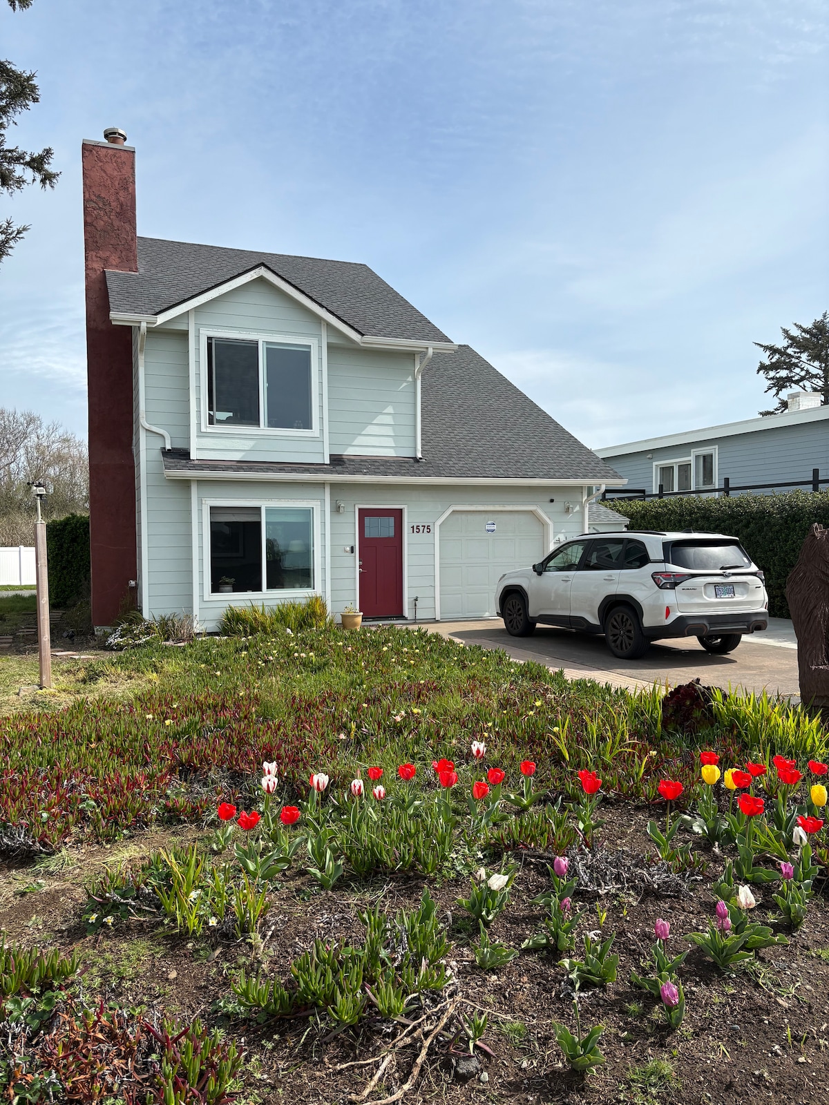 A two-story house features a light blue exterior with a burgundy front door. A vibrant flowerbed of red and pink tulips decorates the front yard. A gray vehicle is parked in the driveway beside the well-maintained lawn.