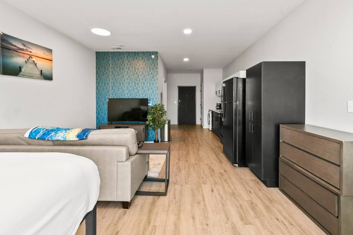 A contemporary studio apartment is showcased with a vibrant blue accent wall and light wood flooring. A sofa sits beside a television and an indoor plant. The kitchen with sleek black appliances is visible, along with a dresser highlighting minimalist design.