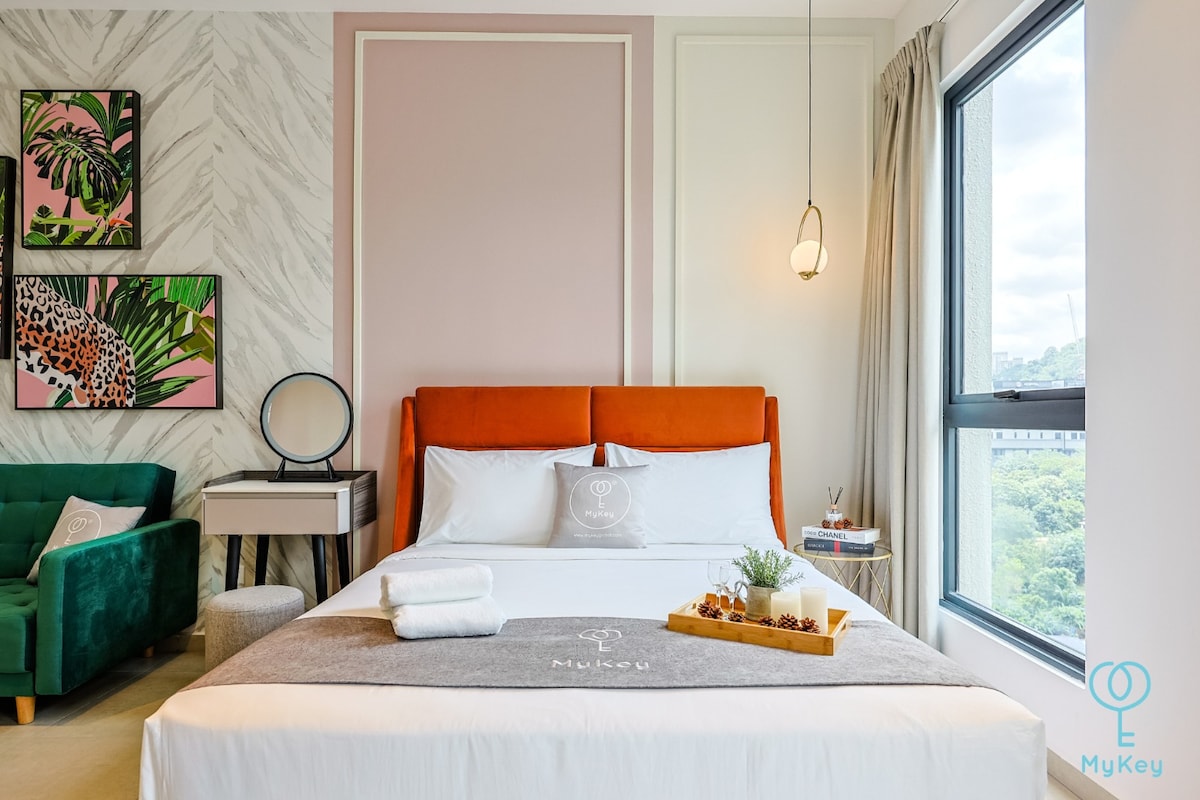 A stylish room features a spacious bed with an orange headboard and crisp white linens. Decorative pillows add a touch of color. A small side table with a mirror is positioned next to a green armchair. Large windows allow natural light to fill the space, highlighting the wall art.