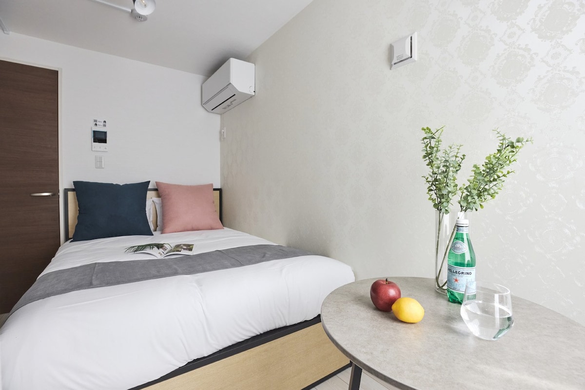 A simple and modern room features a spacious bed with soft bedding, complemented by decorative pillows. A small table holds a glass of water, a green lemon, and a red apple. A potted plant adds a touch of greenery, while an air conditioning unit is mounted on the wall.