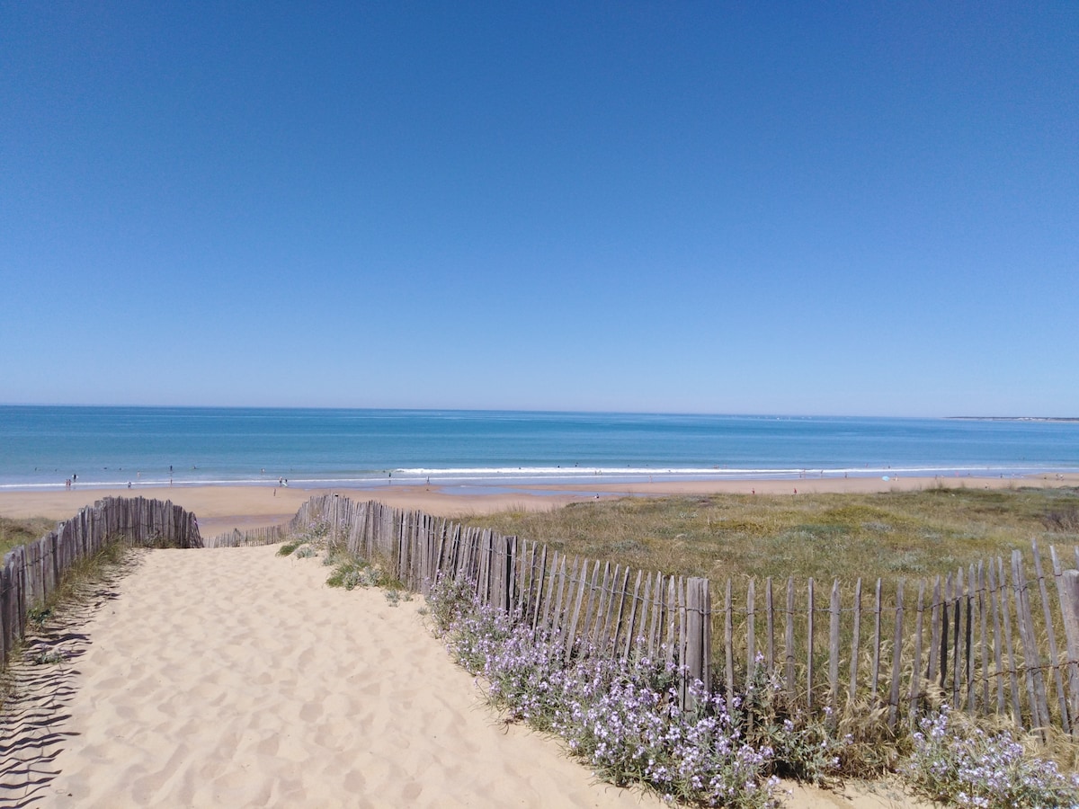 A sandy path leads towards a gentle slope down to a beach, bordered by a wooden fence. The shoreline is visible, with gentle waves lapping at the sand under a clear blue sky. Sparse green grass and purple flowers grow along the path.
