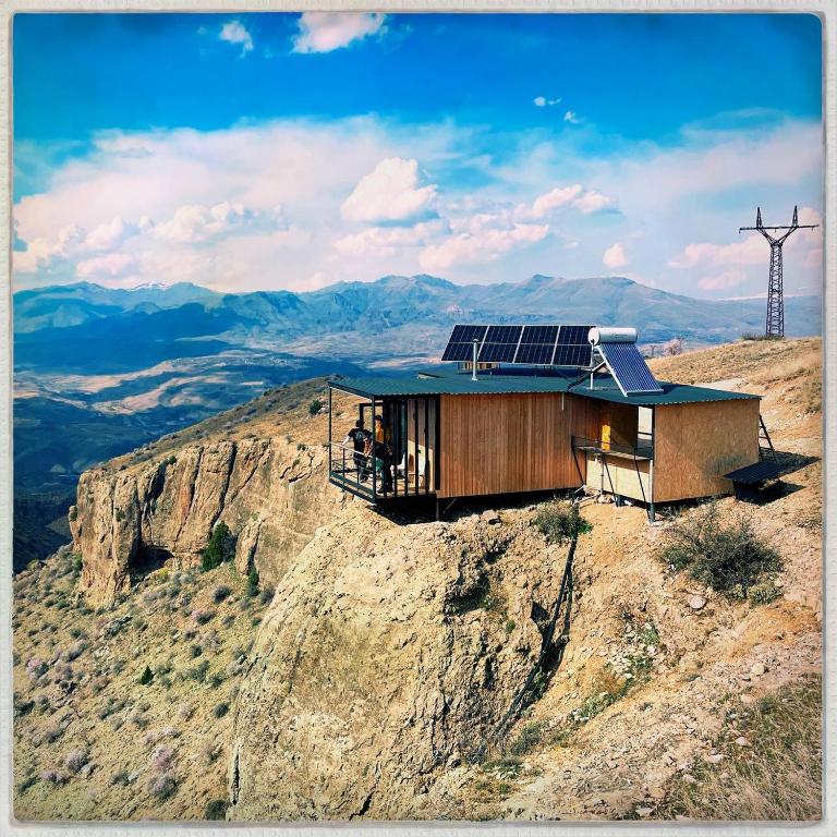 A modern retreat is situated on a rocky cliff, offering sweeping views of the valley and distant mountains. Solar panels are mounted on the roof, and a deck extends outward, providing an outdoor space to enjoy the scenery.