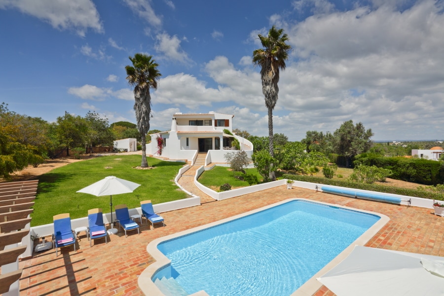The image captures a spacious outdoor area featuring a swimming pool surrounded by sun loungers and a shaded umbrella. Lush greenery and tall palm trees enhance the scene, while a two-story villa is visible in the background against a partly cloudy sky.