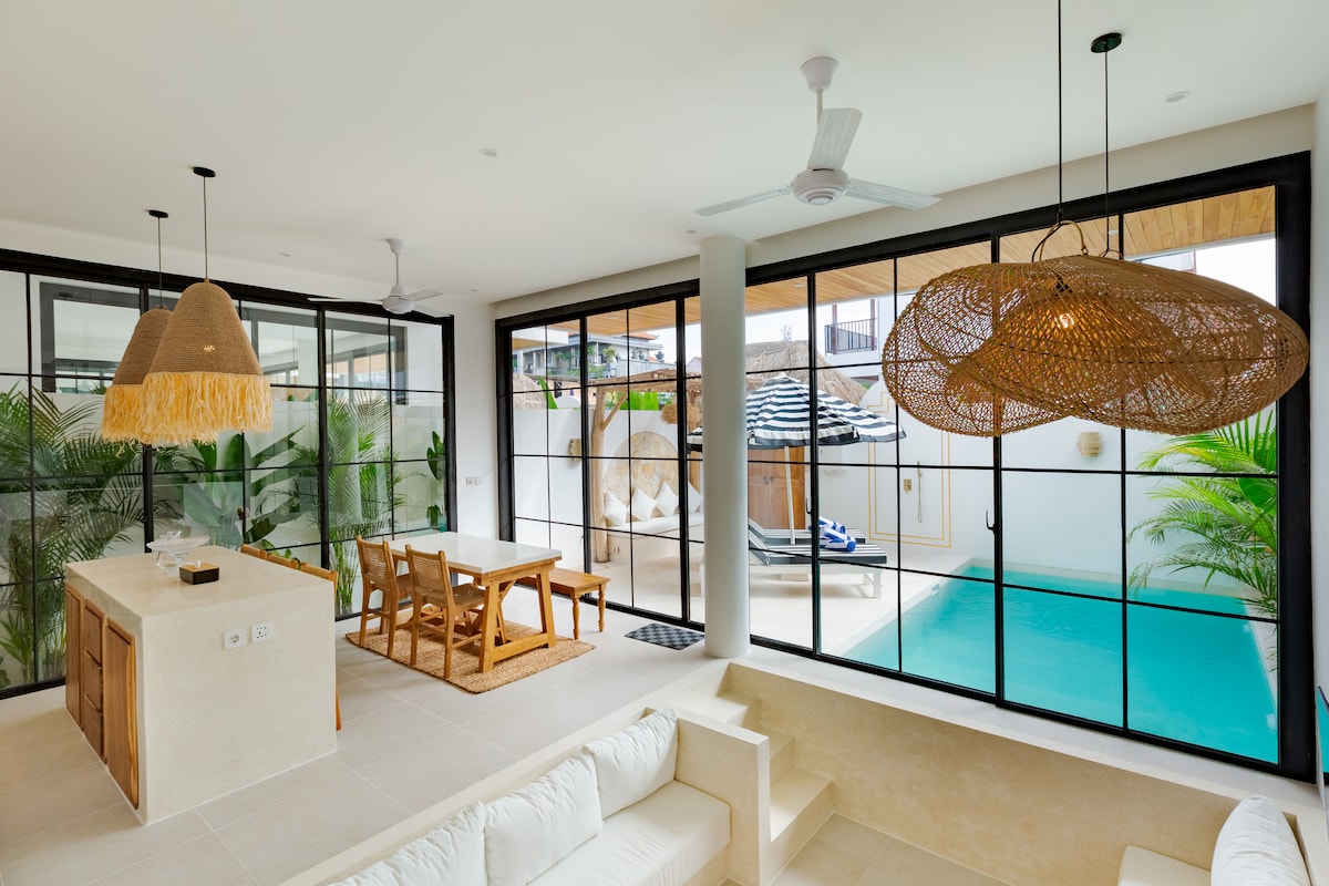A modern living area is presented with tall glass walls, allowing natural light to fill the space. A spacious dining table and kitchen island are visible, while the outdoor pool and lounge area are framed beyond, showcasing tropical plants and a serene atmosphere.
