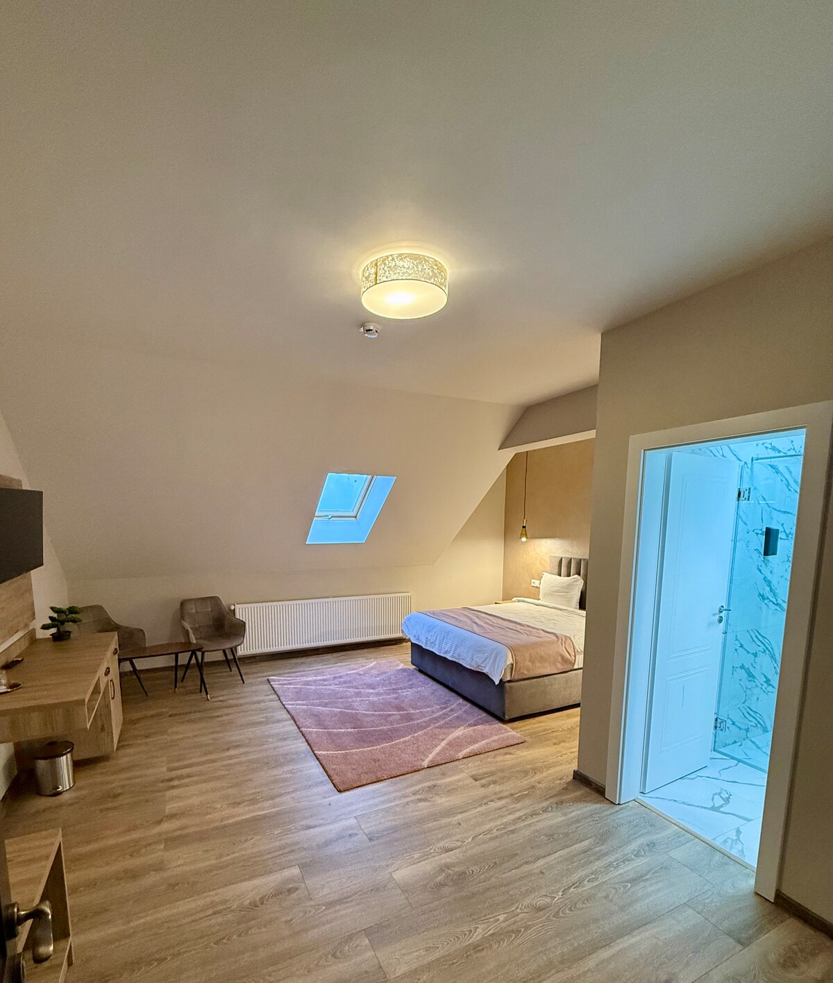 The elegantly arranged room features a queen-size bed with soft linens and a natural light-emitting window. A small seating area with a chair is present, alongside a media console equipped with a TV. The ensuite bathroom is visible through an open door, showcasing modern finishes.