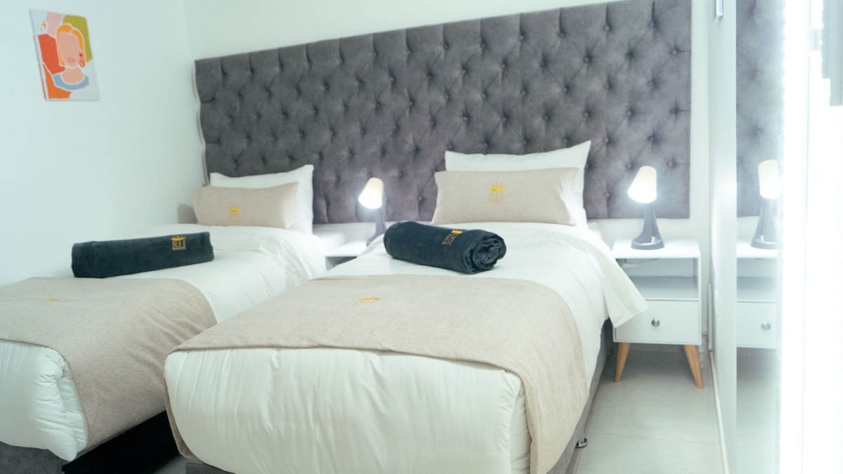 A well-appointed sleeping area is furnished with two spacious beds, each dressed in neutral-toned bedding. Flanking the beds are sleek nightstands topped with modern lamps, providing ample lighting. A plush, tufted headboard in a soft gray serves as a backdrop, adding texture to the room.